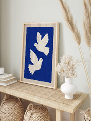Two white illustrative doves flying against a deep blue background with a beige border, in an alternate framed mockup