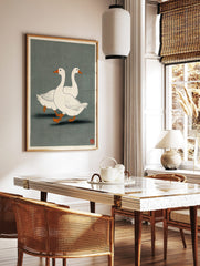 Illustrative print of two white geese with orange beaks and feet walking on a dark grey-green, in a styled room mockup