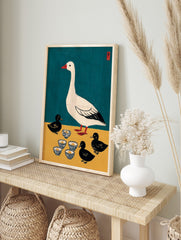 Illustrative print of a white goose and three black ducklings with patterned bowls on teal, in an alternate framed mockup