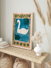 White swan with yellow beak swimming in blue water with green reeds and an ornate botanical, in an alternate framed mockup