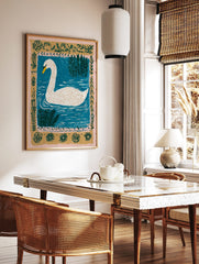 White swan with yellow beak swimming in blue water with green reeds and an ornate botanical border, in a styled room mockup