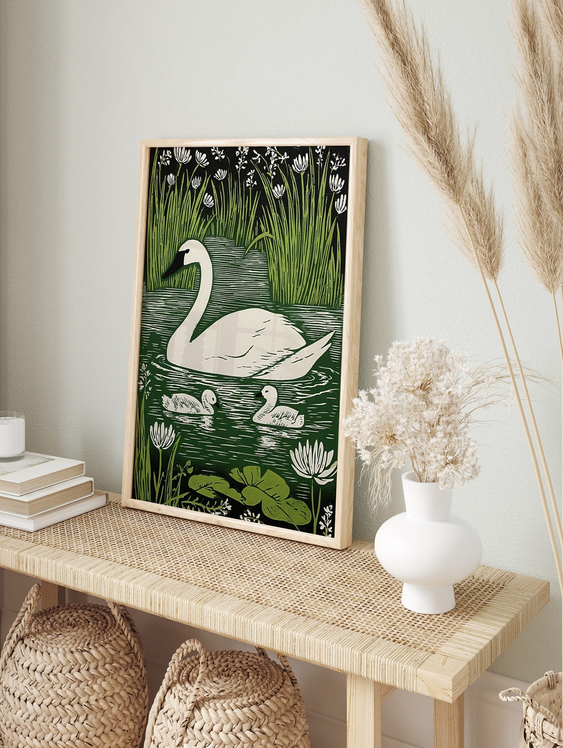 White swan and two cygnets on green water with reeds and white flowers in linocut print style, in an alternate framed mockup