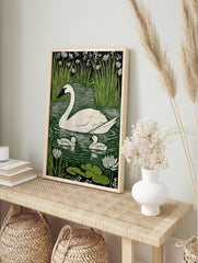 White swan and two cygnets on green water with reeds and white flowers in linocut print style, in an alternate framed mockup