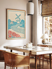 Illustrative windmill landscape art print with a bridge, cottage, and red flowers in a blue and, in a styled room mockup
