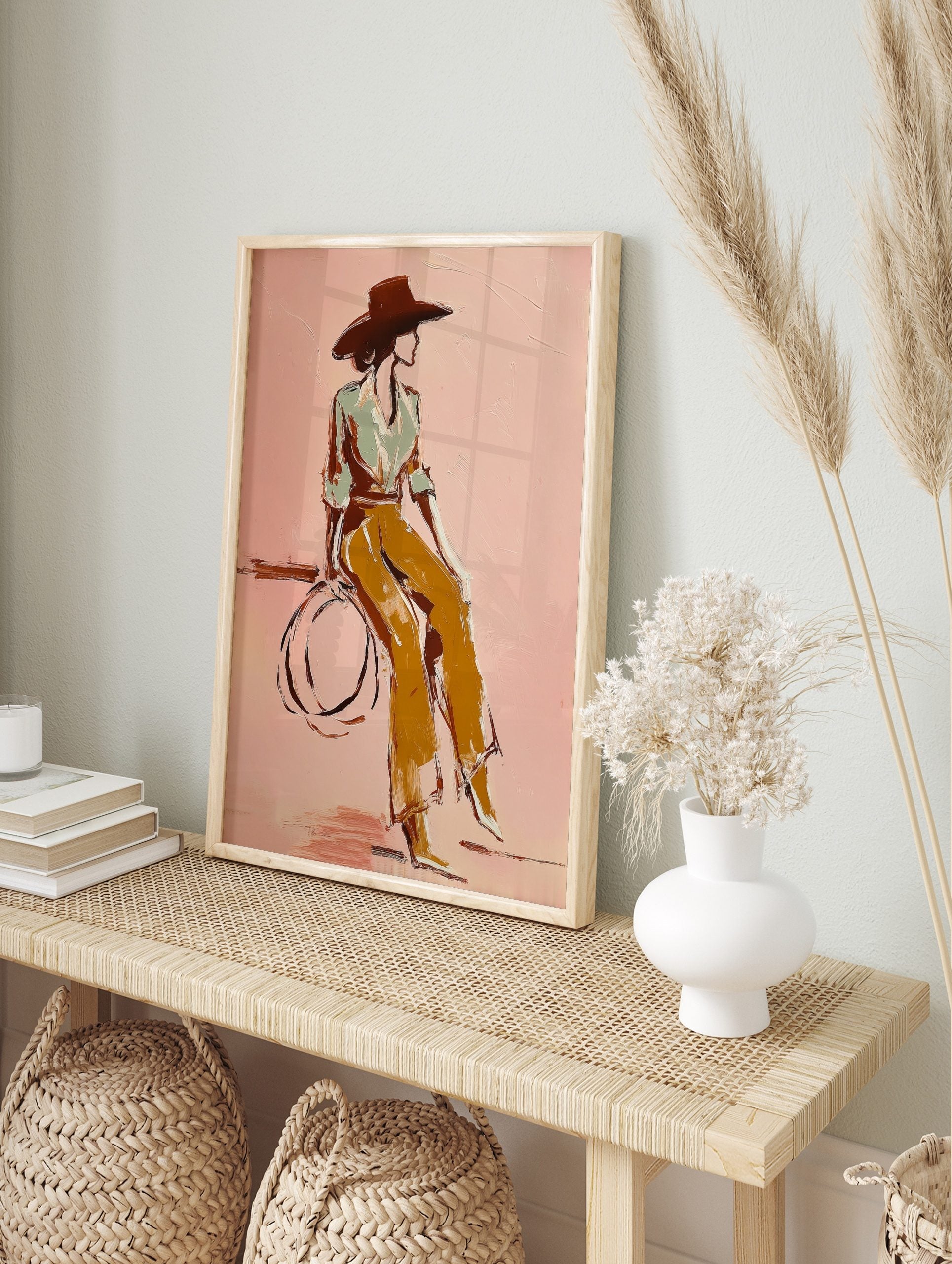 Illustrative print of woman in wide hat and ochre trousers against a pink background, in an alternate framed mockup