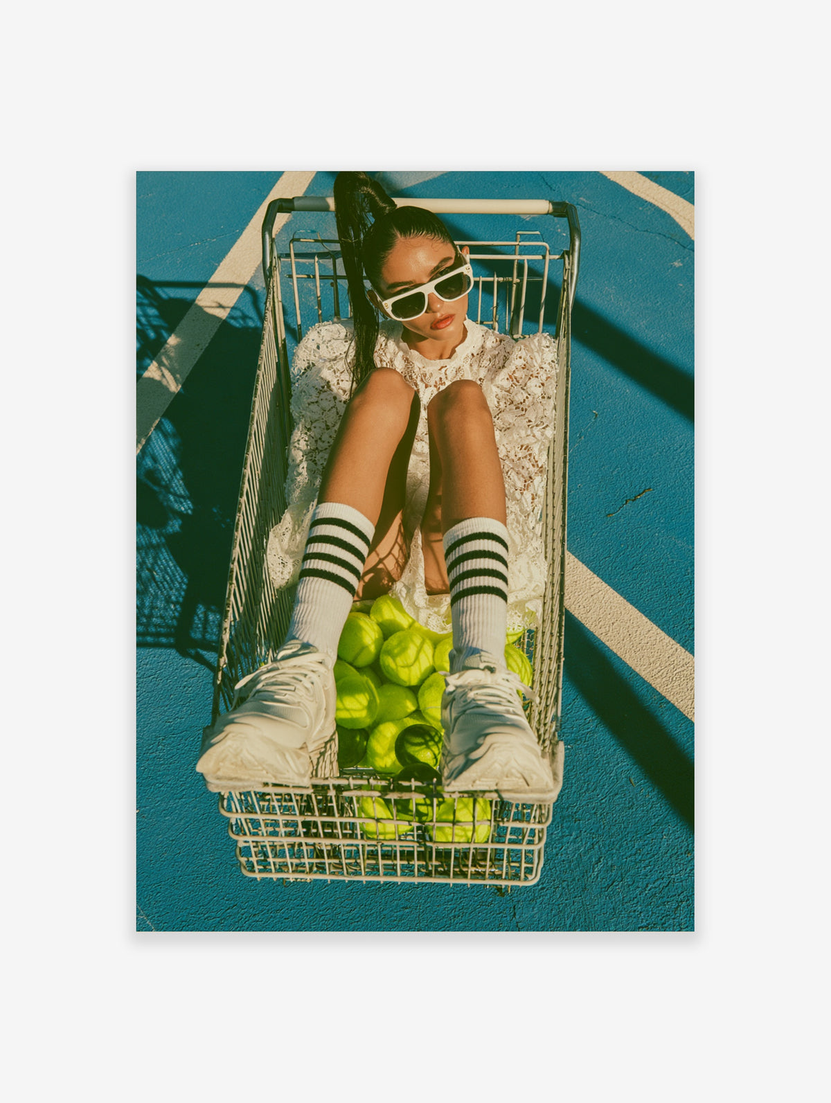 Woman in a shopping cart with tennis balls on a blue court, wearing white lace and sunglasses