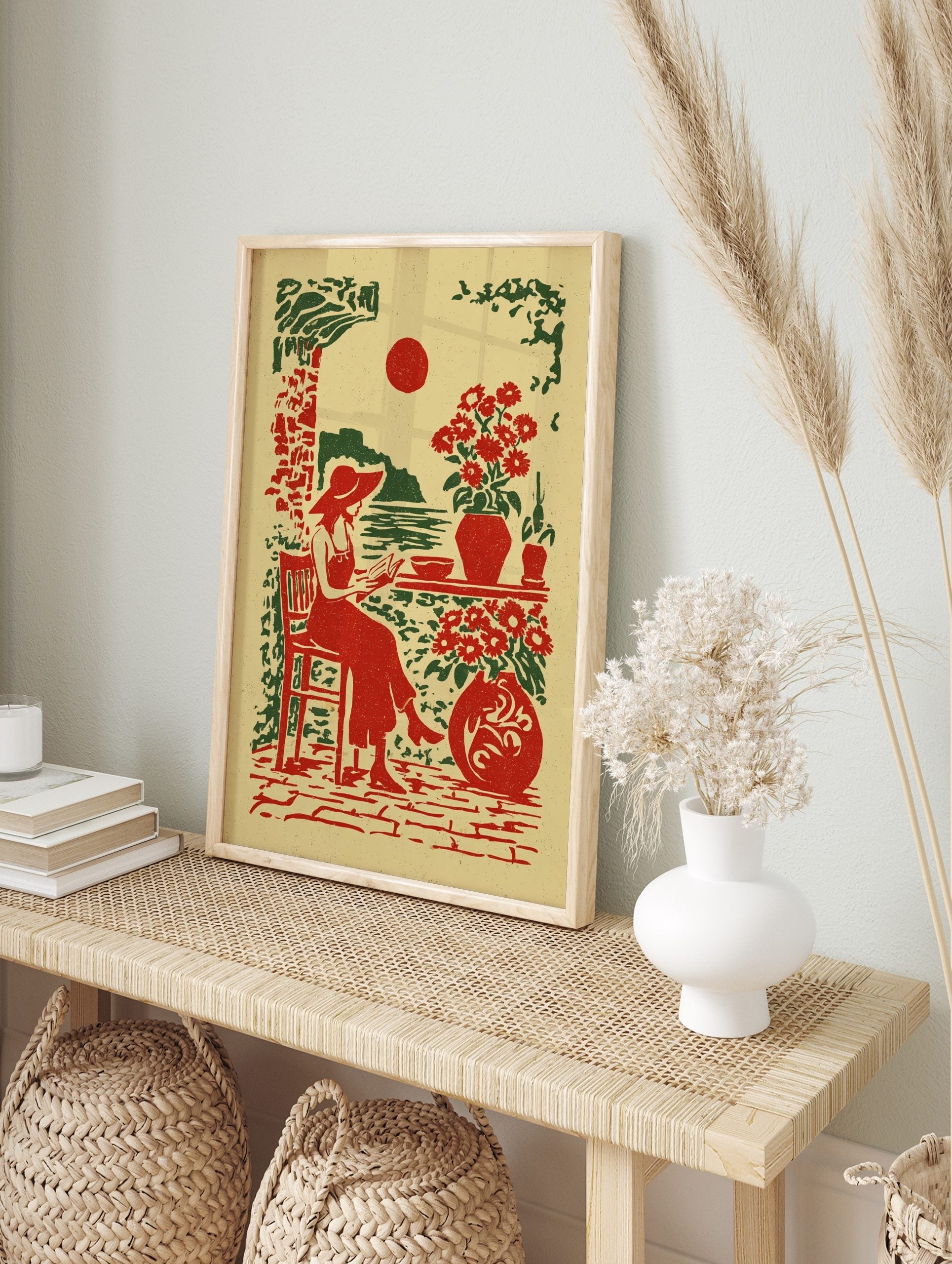 Illustrative print of a woman in a hat reading by the sea with red sun and potted flowers in, in an alternate framed mockup