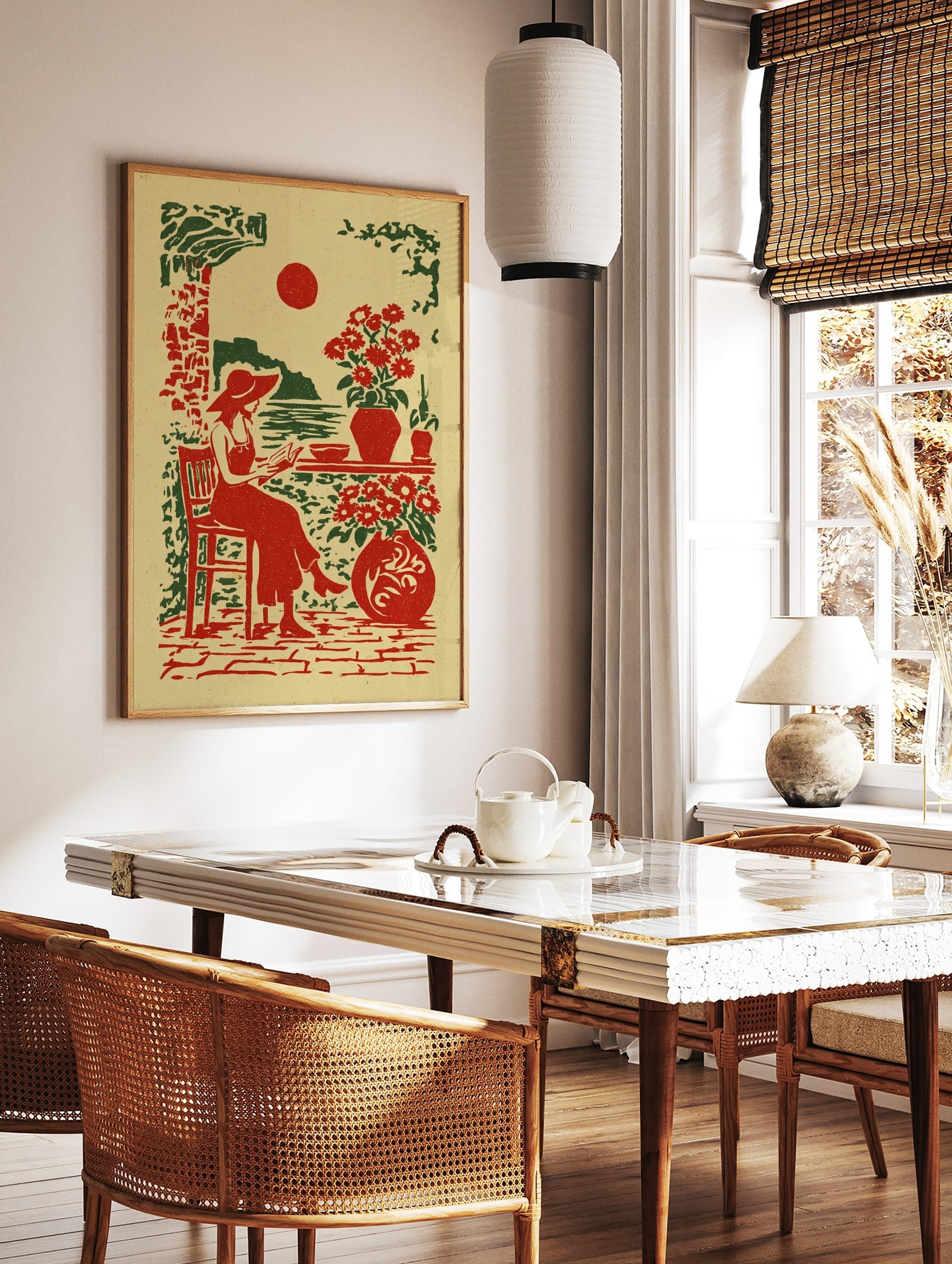 Illustrative print of a woman in a hat reading by the sea with red sun and potted flowers in red, in a styled room mockup