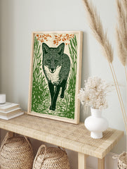Illustrative woodcut-style art print of a green fox walking through grass with orange leaves, in an alternate framed mockup