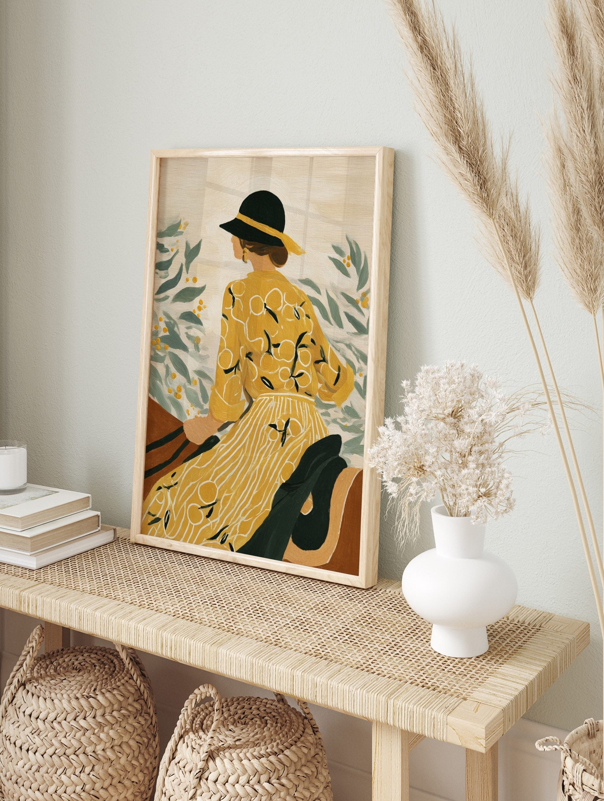Illustrative art print of a woman in yellow dress and black hat, looking at green leaves with, in an alternate framed mockup