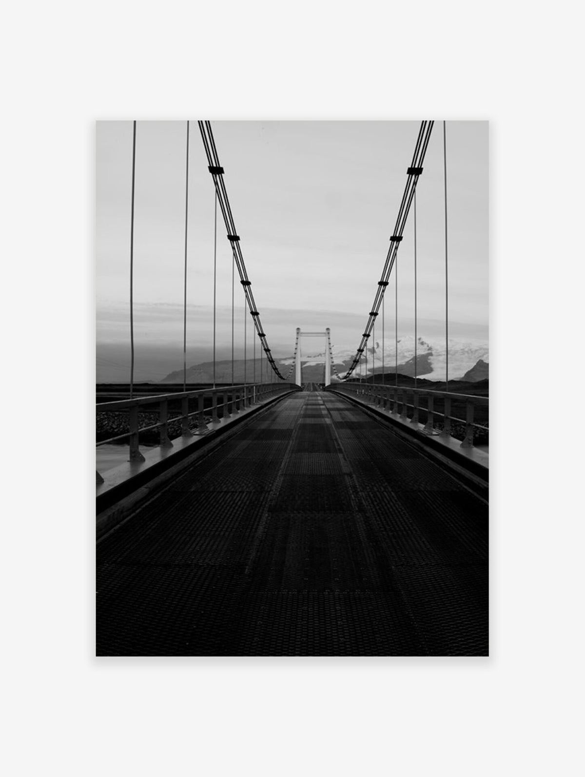 Vertical black and white photograph of a suspension bridge leading toward a distant mountain range.