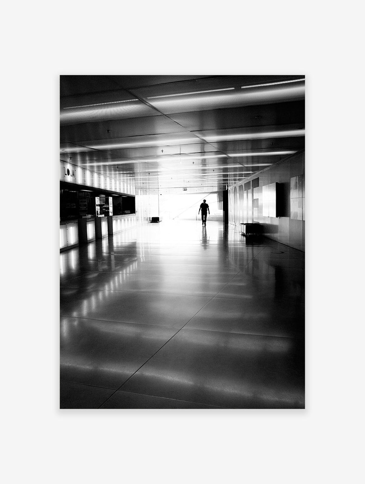 A black and white photograph of a person walking down a long, brightly lit corridor with reflective floors.