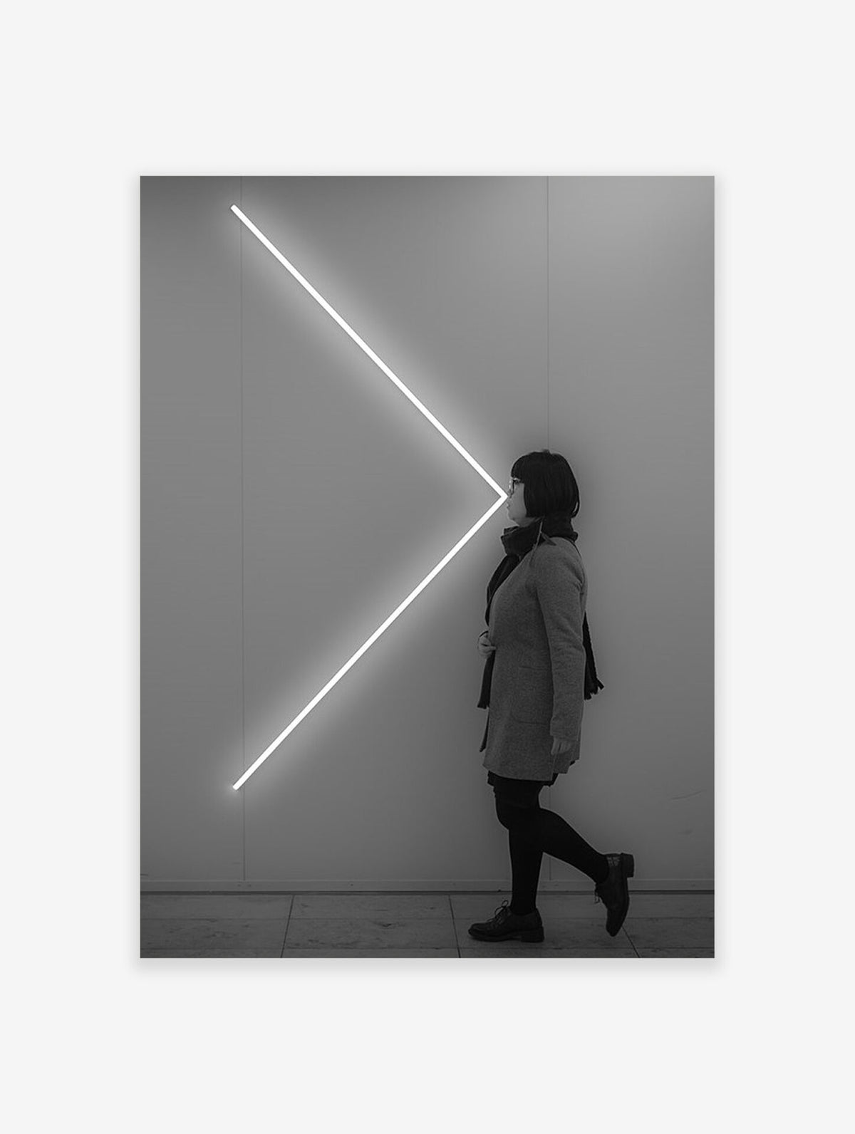 Black and white photograph of a woman walking past a glowing white V-shaped neon light on a wall.
