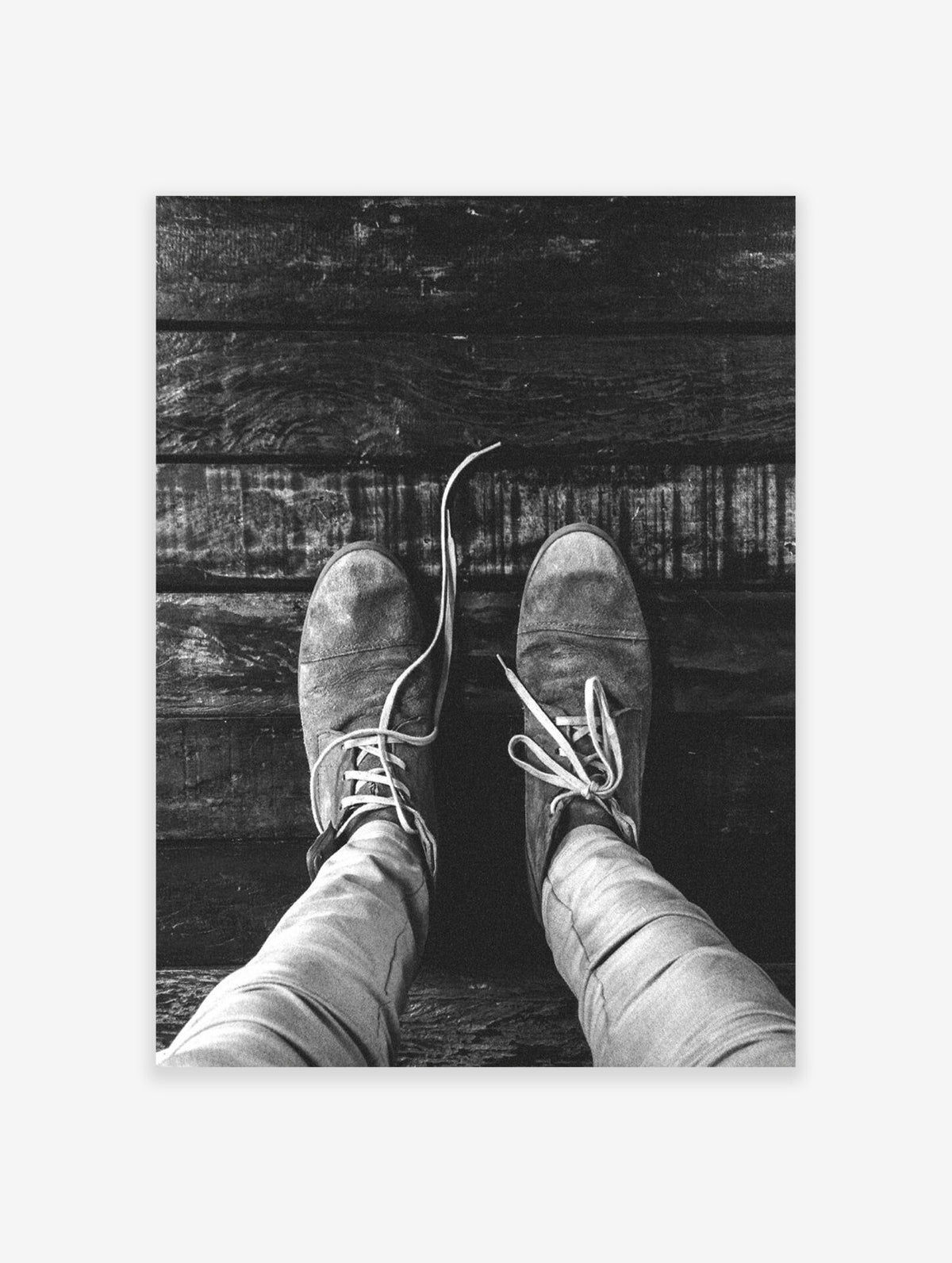 Black and white close-up of a person's feet in laced shoes resting on wooden planks.